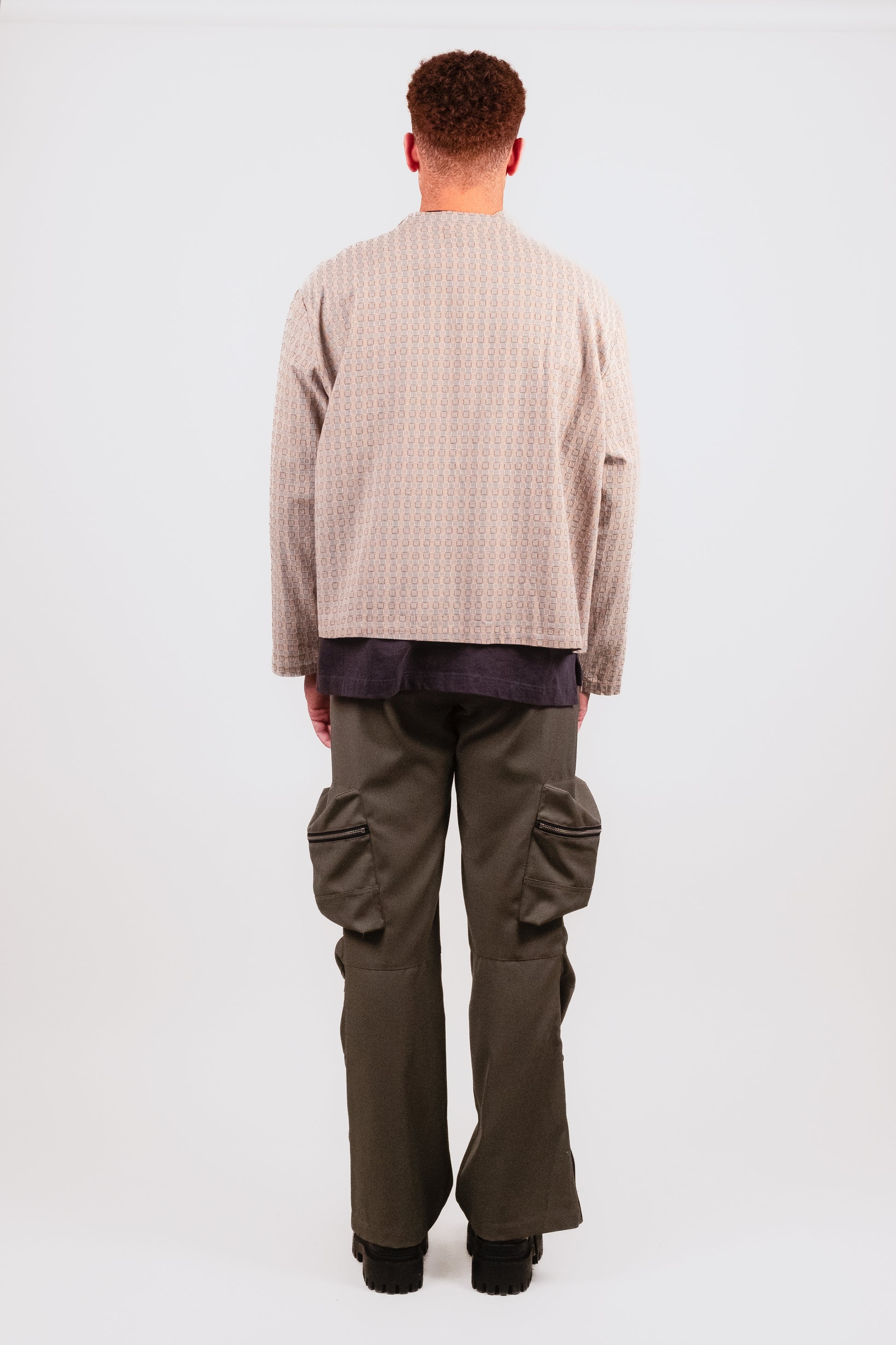 Male model back view in Technicalist Cargo — minimalist suiting-style trousers with flare-zip ankle and single zip back pocket - and Smart Zip — smart, trim, cropped and boxy jacket with beige and blue woven pattern and minimal finishing