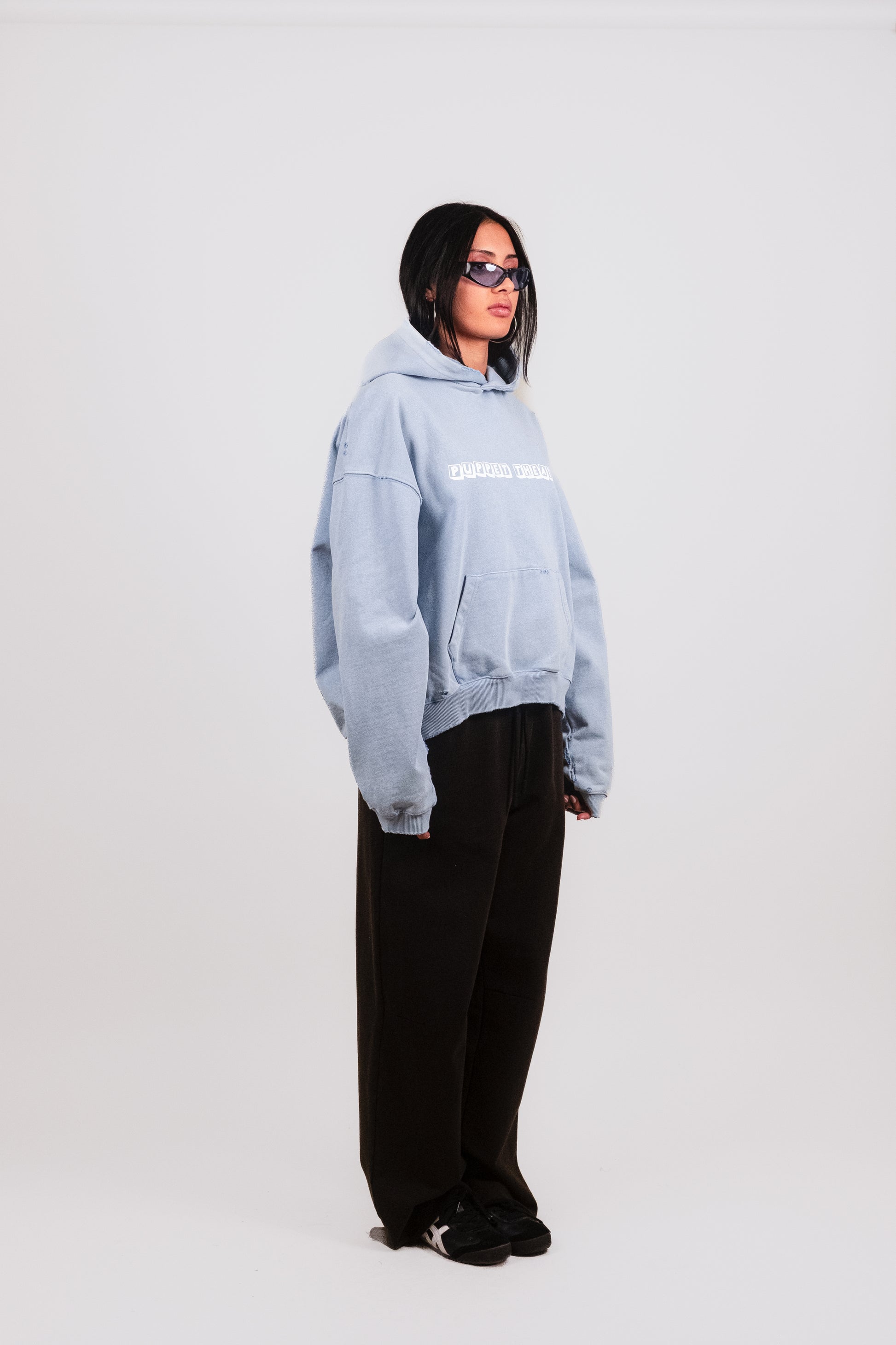 Anisa in alternate 1/4 view styling the Reminiscence Block Logo Hoodie — pastel baby blue luxury hoodie with cropped silhouette, raw hems, faded hand-finished treatment, and iconic graphic branding inspired by the defunct Denver Puppet Theater.
